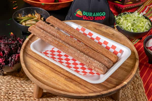 Freshly made churros coated in cinnamon sugar, served on a red checkered paper-lined tray with traditional Mexican sides like hibiscus and sautéed peppers in the background.