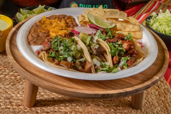 Mexican taco trio with carne asada, carnitas, and al pastor, topped with chopped onions, cilantro, and salsa verde, served alongside cheesy refried beans, tortilla chips, lime, and sliced radish. Set on a rustic wooden plate with festive background elements.