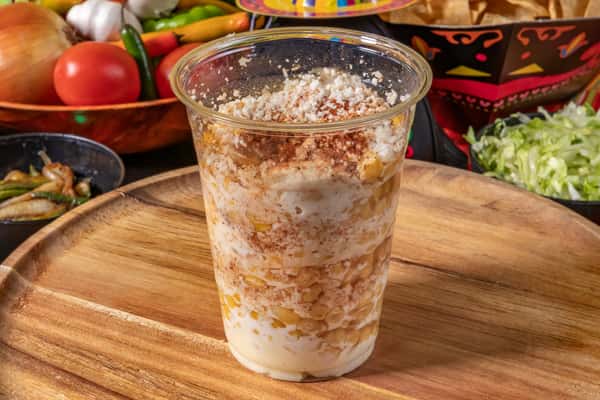 A tall cup of crema-dressed elote-like corn snack dusted with cheese and chili powder, placed near fresh ingredients and sauces; alongside a cheesy enchilada smothered in red sauce and garnished with cheese.