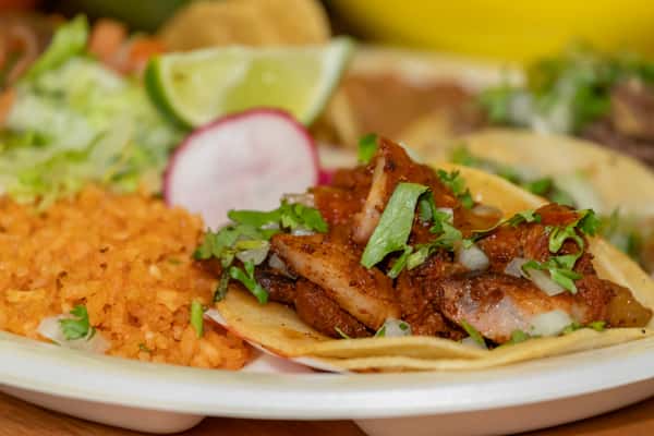 Authentic Mexican taco plate featuring a grilled taco al pastor topped with cilantro and onions, served with seasoned rice, fresh radish, lime, and lettuce.