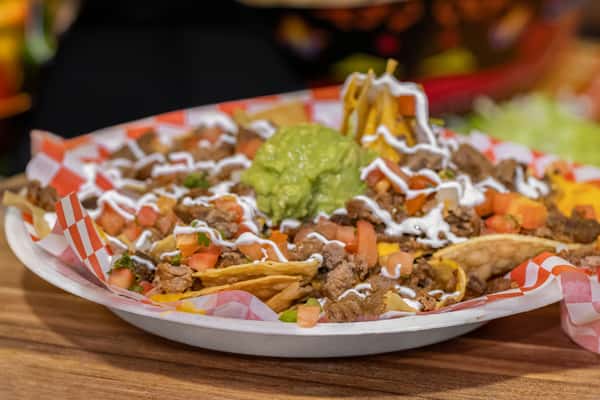 Loaded carne asada nachos topped with melted cheese, diced tomatoes, drizzled crema, and a generous scoop of guacamole, served on a red checkered tray.