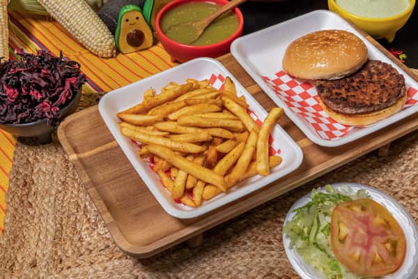 Grilled beef patty on a sesame seed bun served with a side of seasoned French fries and a fresh salad of shredded lettuce and tomato. Displayed with traditional Mexican condiments including green salsa and creamy dressing for a Mexican-American fusion meal.