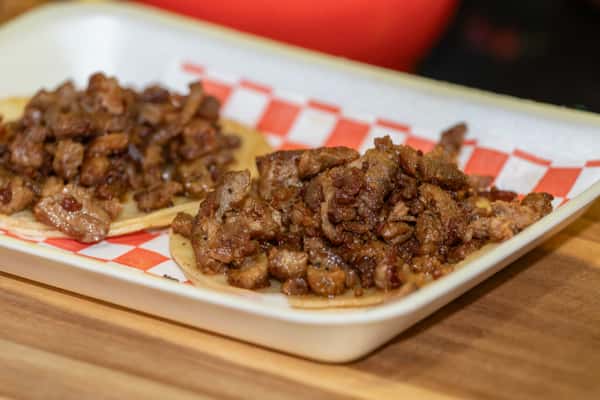 Grilled carne asada tacos on corn tortillas, loaded with tender marinated beef pieces, served on a red checkered paper in a white tray.