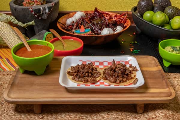 Two carne asada street tacos served on corn tortillas, piled with grilled and seasoned beef. Presented on a red checkered tray alongside traditional Mexican condiments—red salsa, green salsa, guacamole—and surrounded by dried chiles, garlic, avocados, and limes for an authentic taqueria ambiance.