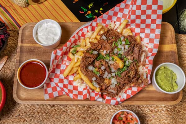 Carne asada fries loaded with shredded beef, chopped onions, melted cheese, and cilantro, served with sides of guacamole, sour cream, and salsa on a wooden tray.