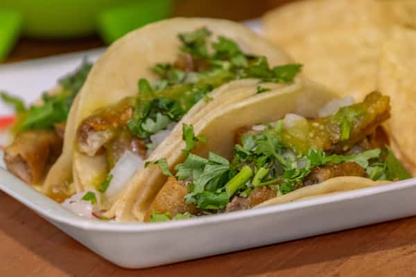Authentic Mexican street tacos filled with crispy pork carnitas, topped with chopped onions, fresh cilantro, and green salsa, served in soft corn tortillas with a side of tortilla chips.