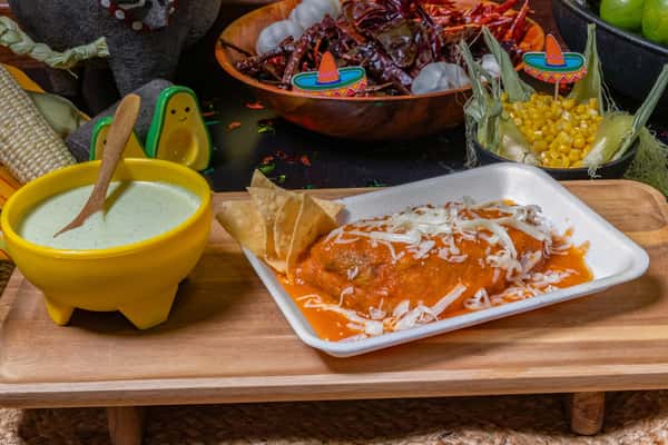 Mexican chile relleno topped with red salsa and shredded cheese, served with crispy tortilla chips and a creamy green sauce, presented on a wooden tray with fresh corn and dried chiles in the background.