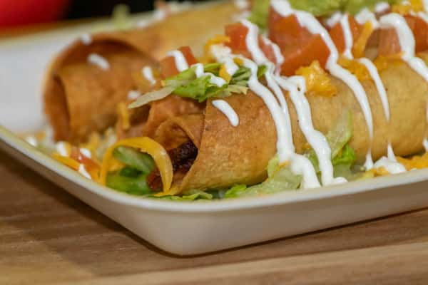 Crispy Mexican taquitos filled with seasoned meat, topped with shredded lettuce, diced tomatoes, cheese, and drizzled sour cream, served on a bed of shredded lettuce.