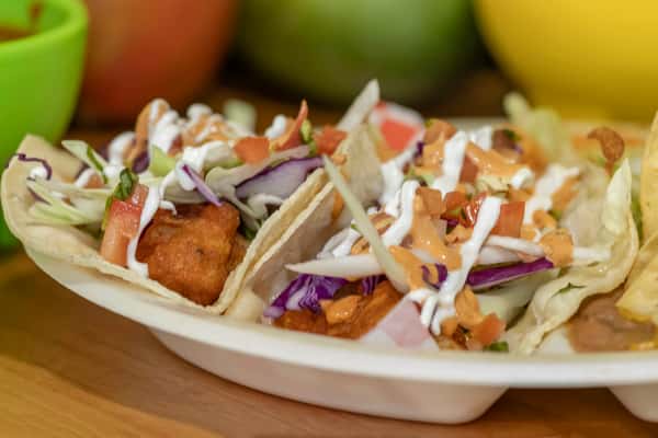 Three fish tacos served on soft tortillas, topped with shredded cabbage, pico de gallo, drizzled creamy sauce, and red sauce; vibrant and fresh seafood taco presentation.