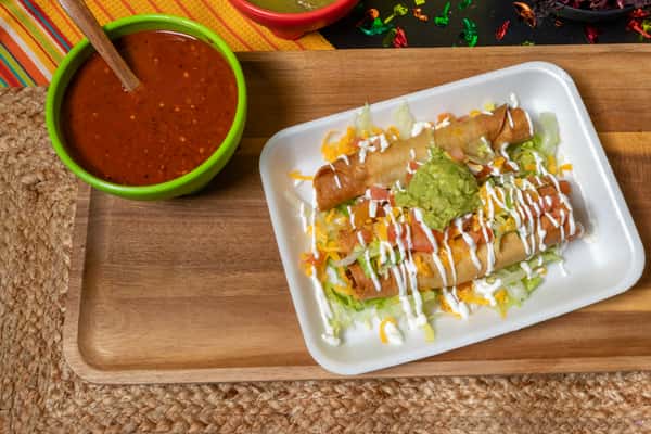 Crispy beef taquitos served on a bed of shredded lettuce, topped with shredded cheese, sour cream drizzle, and guacamole, accompanied by a bowl of red salsa.