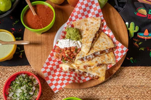 Grilled steak quesadilla stuffed with melted cheese and carne asada, served with guacamole, sour cream, and pico de gallo on a red checkered tray.