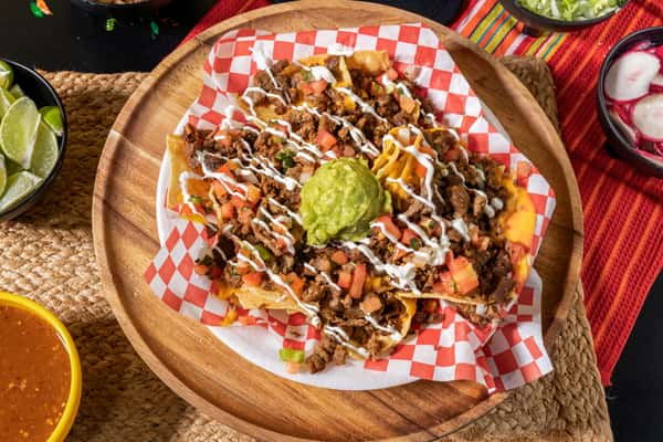 Loaded nachos topped with grilled carne asada, melted cheese, diced tomatoes, sour cream drizzle, and a scoop of guacamole, served on a wooden tray with festive Mexican sides.
