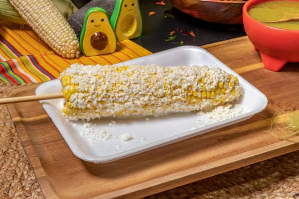 Grilled corn on the cob (elote) coated with crumbled cheese, likely cotija, served on a tray with traditional Mexican condiments in the background.