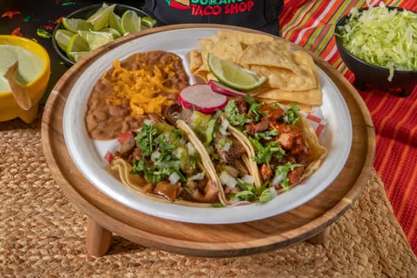 Authentic Mexican street taco plate featuring carne asada, al pastor, and carnitas tacos topped with cilantro, onions, and salsa, served with refried beans, crispy tortilla chips, lime wedges, and sliced radishes. The vibrant setting includes shredded lettuce and a creamy green salsa, capturing a festive taqueria vibe.