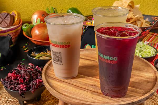 Trio of traditional Mexican drinks featuring horchata, tamarindo, and hibiscus agua fresca, served in clear plastic cups with the Durango Taco Shop logo, surrounded by fresh ingredients.