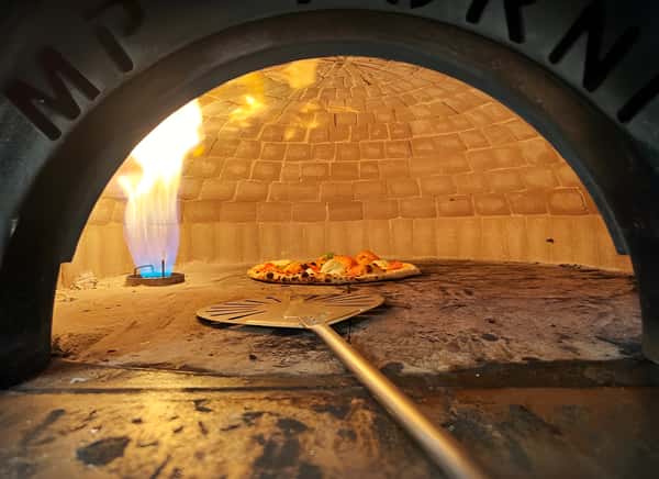 Pizza baking in a brick oven with a blue gas flame visible. A metal pizza peel rests in the foreground.
