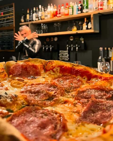 A large pepperoni pizza with melted cheese and red sauce in the foreground, with a blurry bartender in the background.