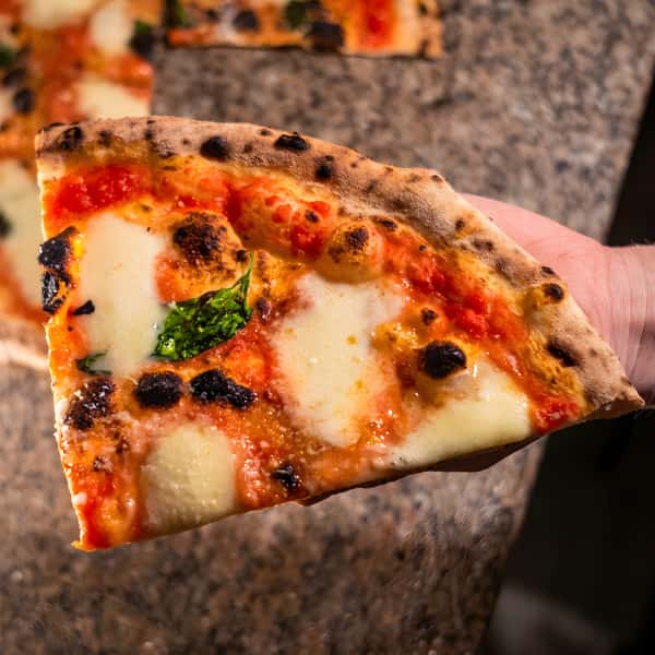 Hand holding a slice of Neapolitan pizza with melted mozzarella, red sauce, and basil.