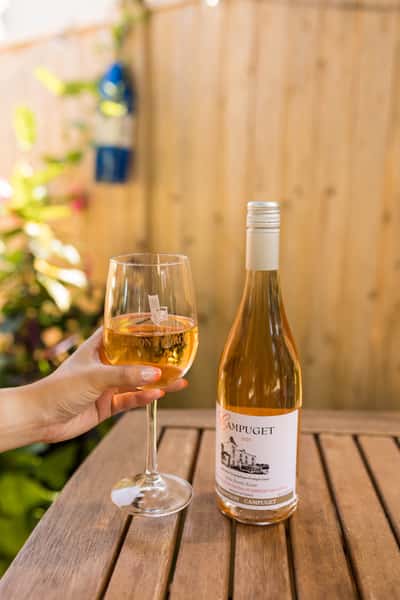 A hand holds a glass of white wine next to a bottle of Campuget wine on a wooden table in a sunny garden setting.