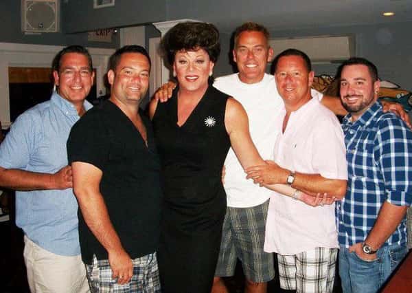 Six people posing closely together at a gathering, with one in a black dress and styled hair, surrounded by smiling men.