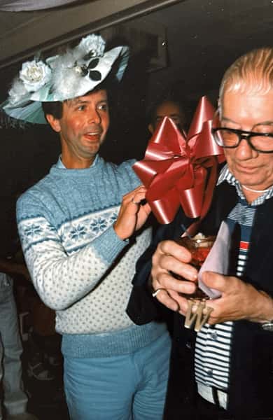 Man in a festive hat smiles while pinning a large red bow on another man holding a cocktail and looking down.