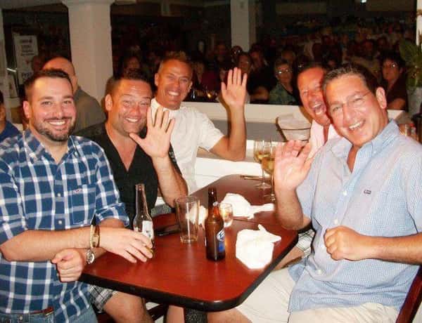 Six men smiling and waving at a crowded bar table, drinks in hand, with a lively audience in the background.