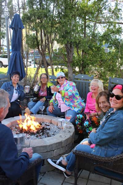 A cheerful group of friends gathered around a cozy outdoor fire pit, enjoying drinks and lively conversation on a sunny day.