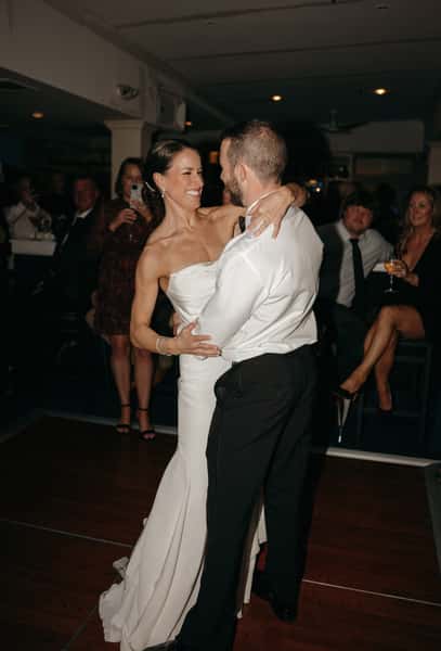 Bride and groom sharing their first dance on the dance floor, smiling joyfully as guests look on and take photos.