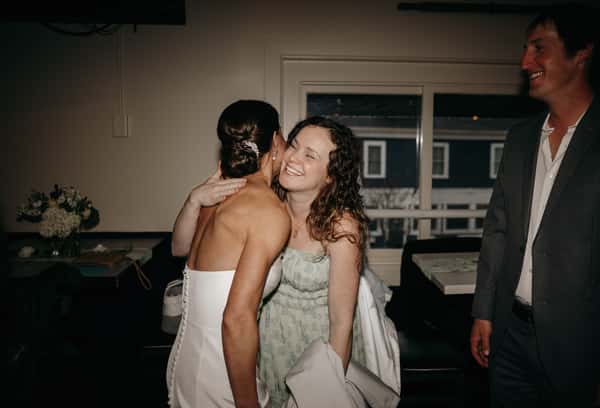 Bride in white dress hugs smiling guest in mint green dress, man in suit watches, during a joyful wedding reception indoors.