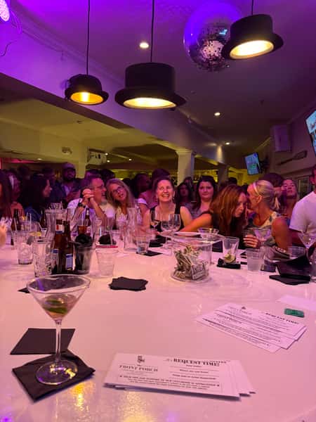 Crowded bar scene with people enjoying drinks under purple lighting, a disco ball, and hat-shaped light fixtures.