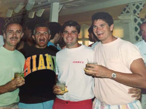 Four men smiling and holding drinks at a lively indoor gathering, one wearing a "MIAMI" shirt and another a "JIMMYZ" tee.