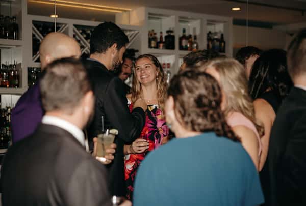 Smiling woman in a floral dress talking with guests at a crowded bar during an evening celebration.