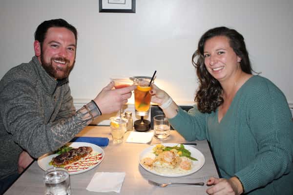 Smiling couple toasting cocktails at a restaurant table with shrimp risotto and seared tuna entrées.