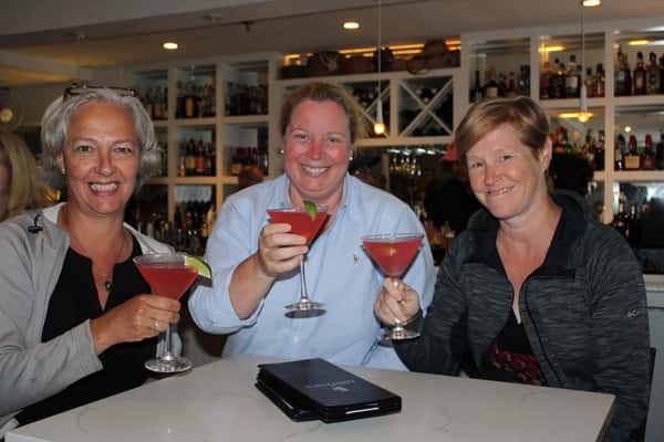 Three smiling women holding pink cocktails with lime wedges, seated at a bar table with menus in front of a mirrored liquor shelf.