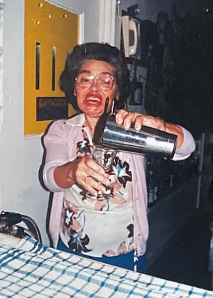 Smiling woman behind a bar pouring a drink from a cocktail shaker into a glass, wearing a floral blouse and pink cardigan.