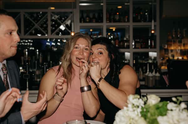 Two women at a bar laughing and eating oysters, surrounded by drinks and floral table decor.