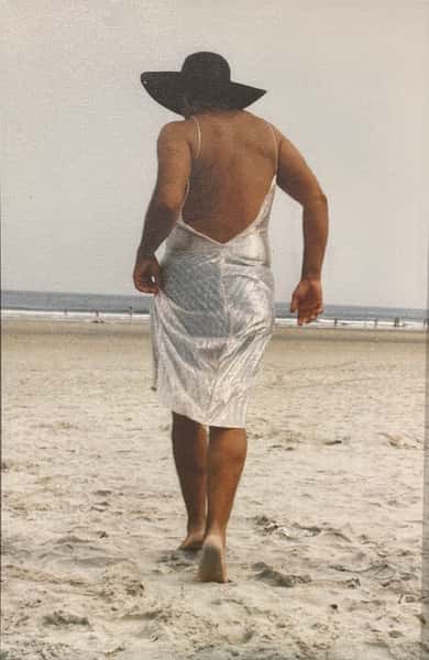 Person wearing a black sunhat and sheer white dress walking barefoot along a sandy beach toward the ocean.