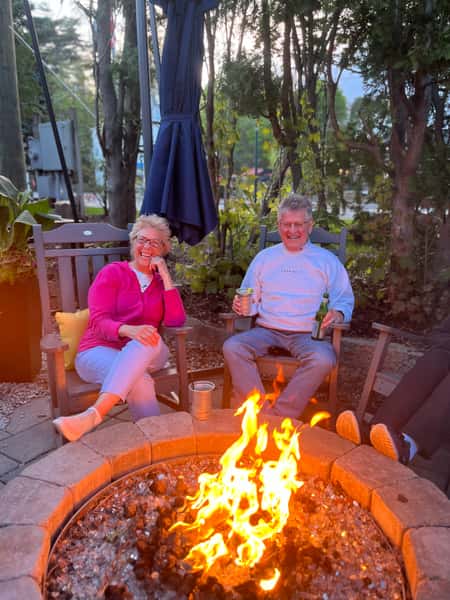 Smiling older couple relaxing with drinks by a fire pit surrounded by trees and evening light.