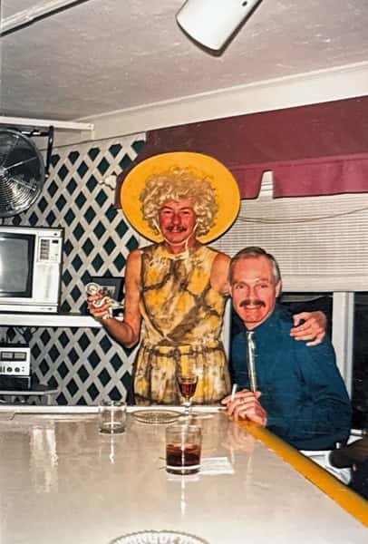 Two men at a bar, one in a blonde wig and yellow hat holding a doll, the other seated with a drink and cigarette.