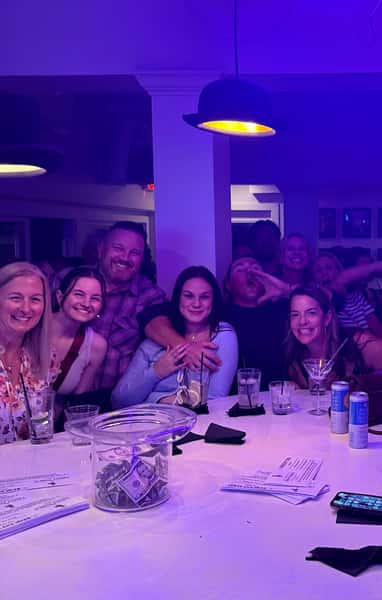 Group of friends smiling and enjoying drinks around a bar table under purple lighting with a tip jar and menus in front.