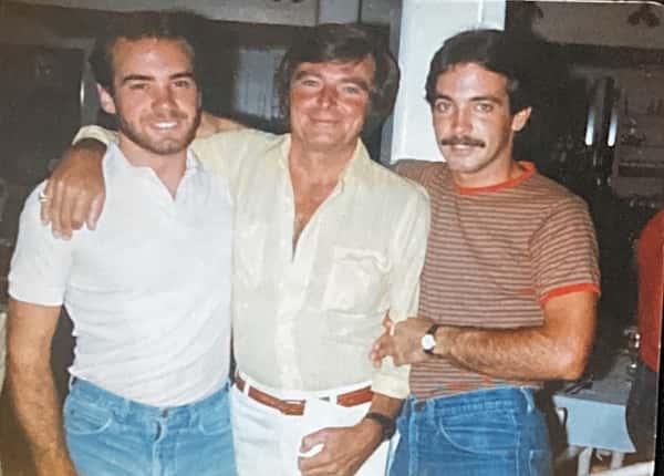Three men smiling and posing closely together indoors, dressed in casual vintage clothing with arms around each other.