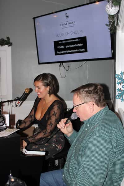 This image captures a live music moment at The Front Porch piano bar and restaurant. Julia Chisholm is seen performing at the piano, smiling warmly, while a guest enthusiastically sings into the microphone beside her.