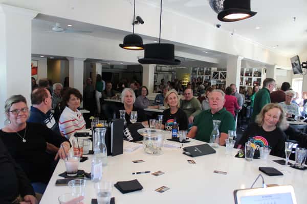 This photo captures a lively, well-attended indoor brunch or lunch gathering, with guests seated around a large circular table. Many are wearing matching "Porch Brunch" shirts, suggesting a themed event or reunion. The setting is bright and sociable, with a bar in the background and laughter filling the room.