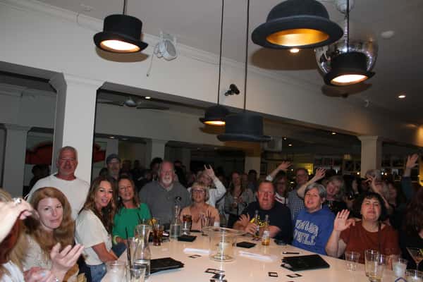A lively indoor gathering with people smiling, waving, and enjoying drinks around a bar-style table. The room features playful top-hat light fixtures and a disco ball, creating a fun and whimsical atmosphere.