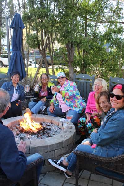 A lively group of people enjoying drinks around an outdoor fire pit. Everyone looks relaxed and cheerful, with trees and a street visible in the background, giving the scene a cozy, festive vibe.