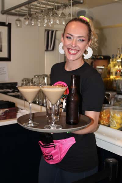 A cheerful server beams brightly while holding a tray with two creamy martinis and a beer, dressed in festive attire and ready to deliver drinks in a lively bar setting.