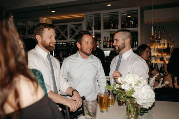 Three men in dress shirts and ties share a laugh at a bar, surrounded by drinks and white floral arrangements.