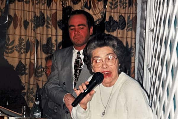 Elderly woman speaking into a microphone beside a man in a suit, standing in front of leafy-patterned curtains.