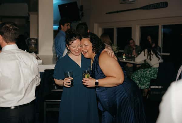 Two women laughing and hugging while holding drinks at a lively indoor celebration, surrounded by other guests enjoying the evening.