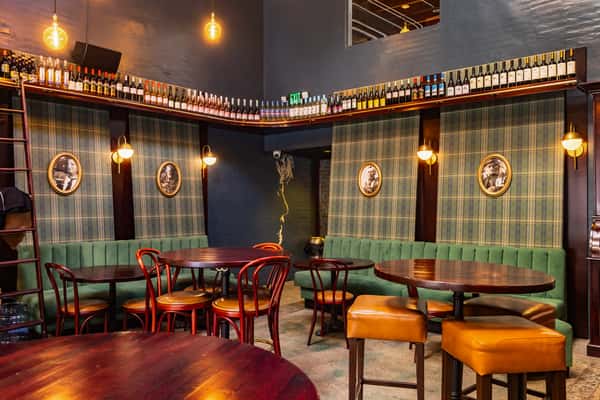 A cozy bar interior featuring green upholstered seating, wooden tables, and vintage portraits on plaid walls, with a shelf of assorted bottles above. Warm lighting enhances the ambiance.